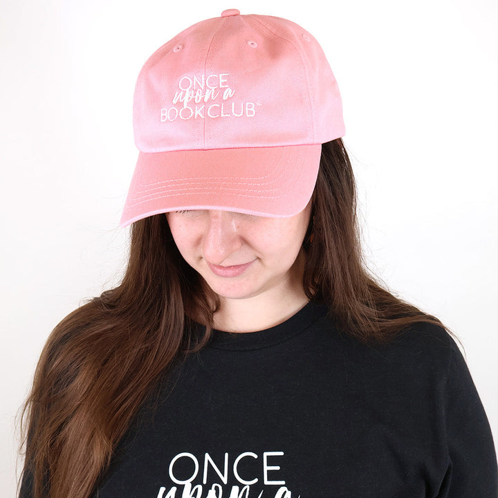 A woman wearing a light pink baseball cap with white embroidered text reading 'Once Upon a Book Club' and a black graphic T-shirt with matching white text, photographed against a plain white background.