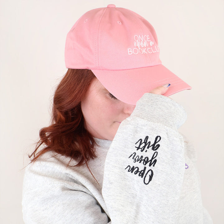 A woman wearing a light pink baseball cap with the words 'Once Upon a Book Club' in white embroidered text and a light gray sweatshirt with black script embroidery on the sleeve that says 'Open Your Gift', posed against a plain off-white background.