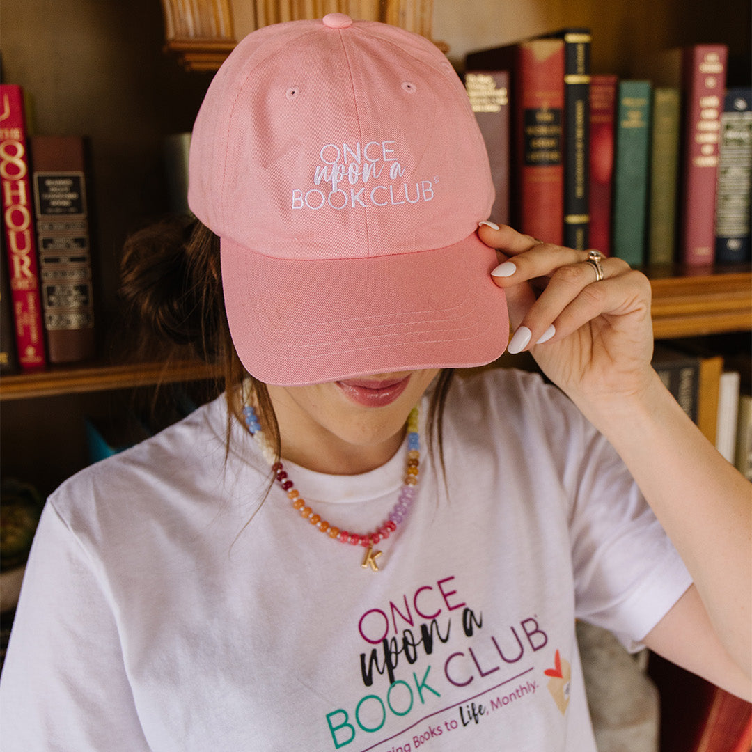 A woman wearing a pink baseball cap and a white graphic T-shirt in front of a bookshelf. The cap and shirt both feature the text 'Once Upon a Book Club' in stylized lettering.