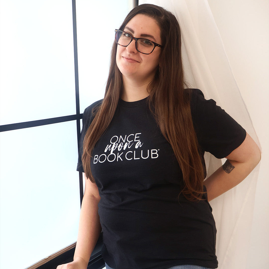 A woman with long brown hair and glasses wearing a black graphic t-shirt that reads 'Once Upon a Book Club' in white script, posing next to a window with white drapes.