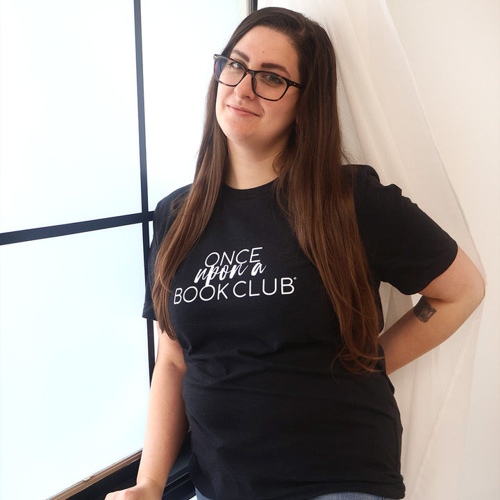 A woman with long brown hair and glasses wearing a black graphic t-shirt that reads 'Once Upon a Book Club' in white script, posing next to a window with white drapes.