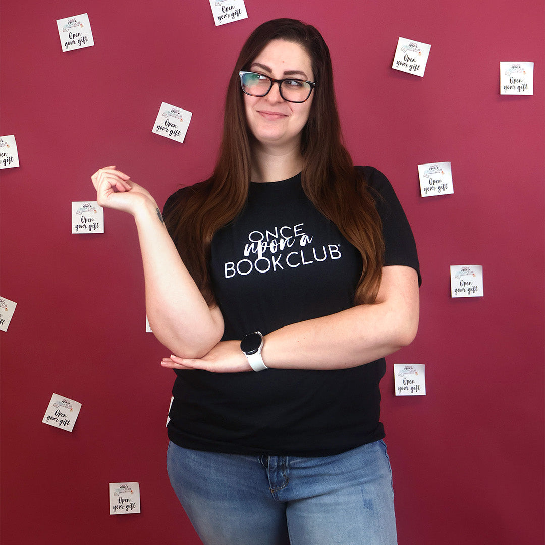 A woman wearing glasses and a black graphic t-shirt that reads 'Once Upon a Book Club' stands against a dark pink wall decorated with small white 'Open Your Gift' sticky notes.