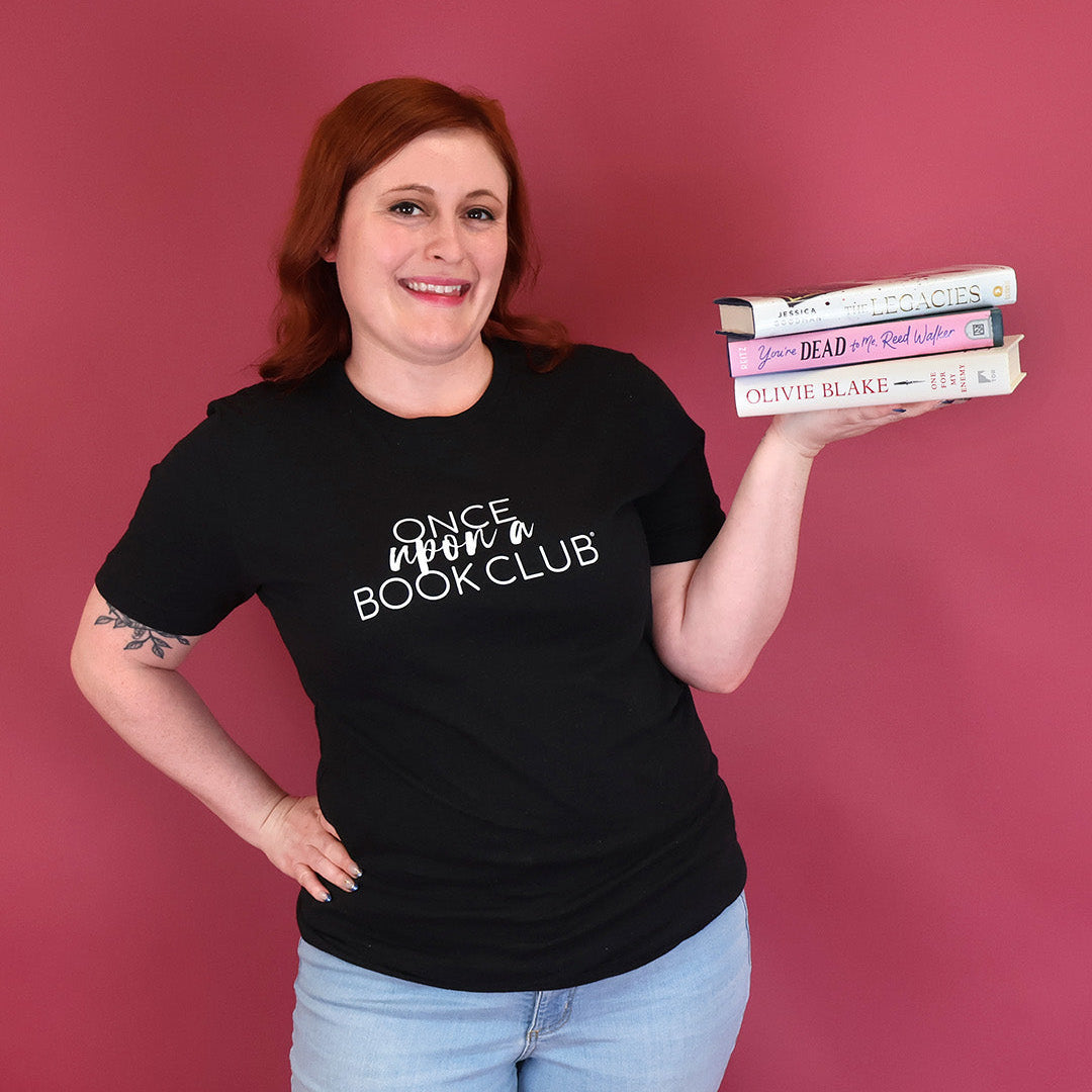 A woman with red hair stands against a solid pink background, wearing a black t-shirt with the white text 'Once Upon a Book Club' and holding a stack of three books.