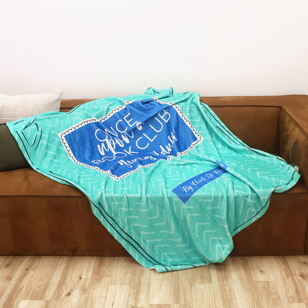 A turquoise and blue patterned throw blanket inspired by the Once Upon a Book Club Young Adult box is draped over a brown leather couch in a living room setting.