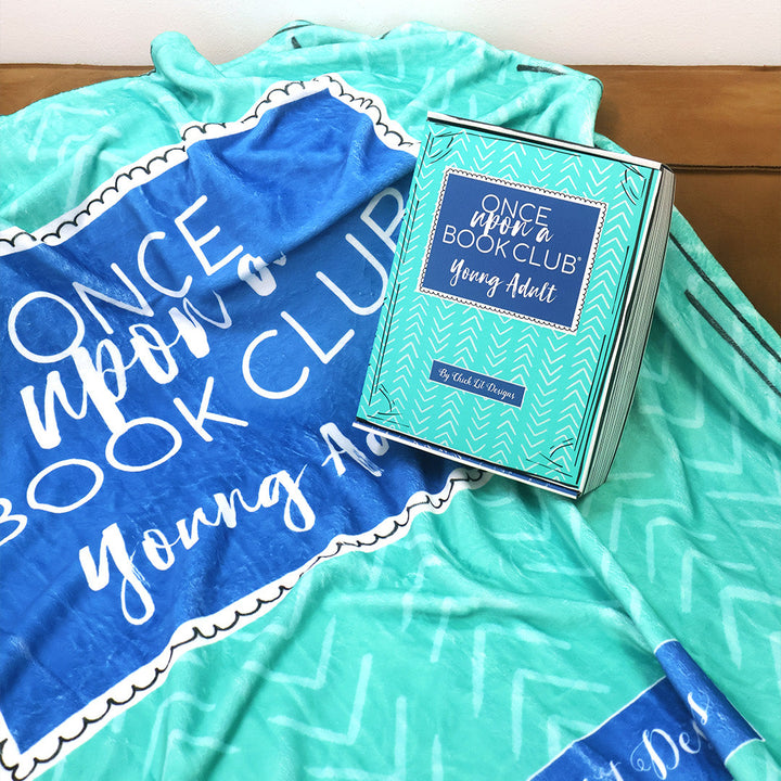 A turquoise and blue patterned throw blanket inspired by the Once Upon a Book Club Young Adult box is draped over a brown leather couch in a living room setting. A matching Young Adult box sits on the blanket on the couch.
