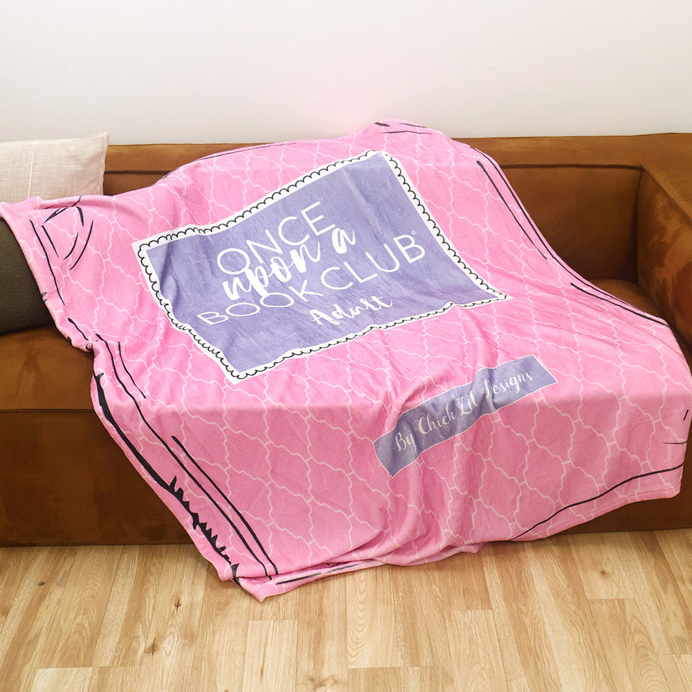 A pink and purple patterned throw blanket inspired by the Once Upon a Book Club Adult box is draped over a brown leather couch in a living room setting.