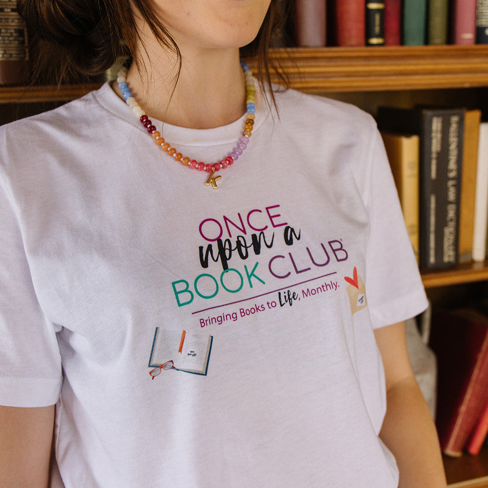 A woman wearing a white graphic t-shirt that reads 'Once Upon a Book Club' in pink, teal, and dark purple lettering set against a bookshelf background.