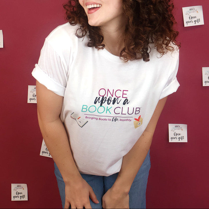 A smiling woman with curly brown hair wearing a white graphic t-shirt that reads 'Once Upon a Book Club' in pink, teal, and dark purple lettering and blue jeans, posing against a dark pink wall decorated with small sticky notes.