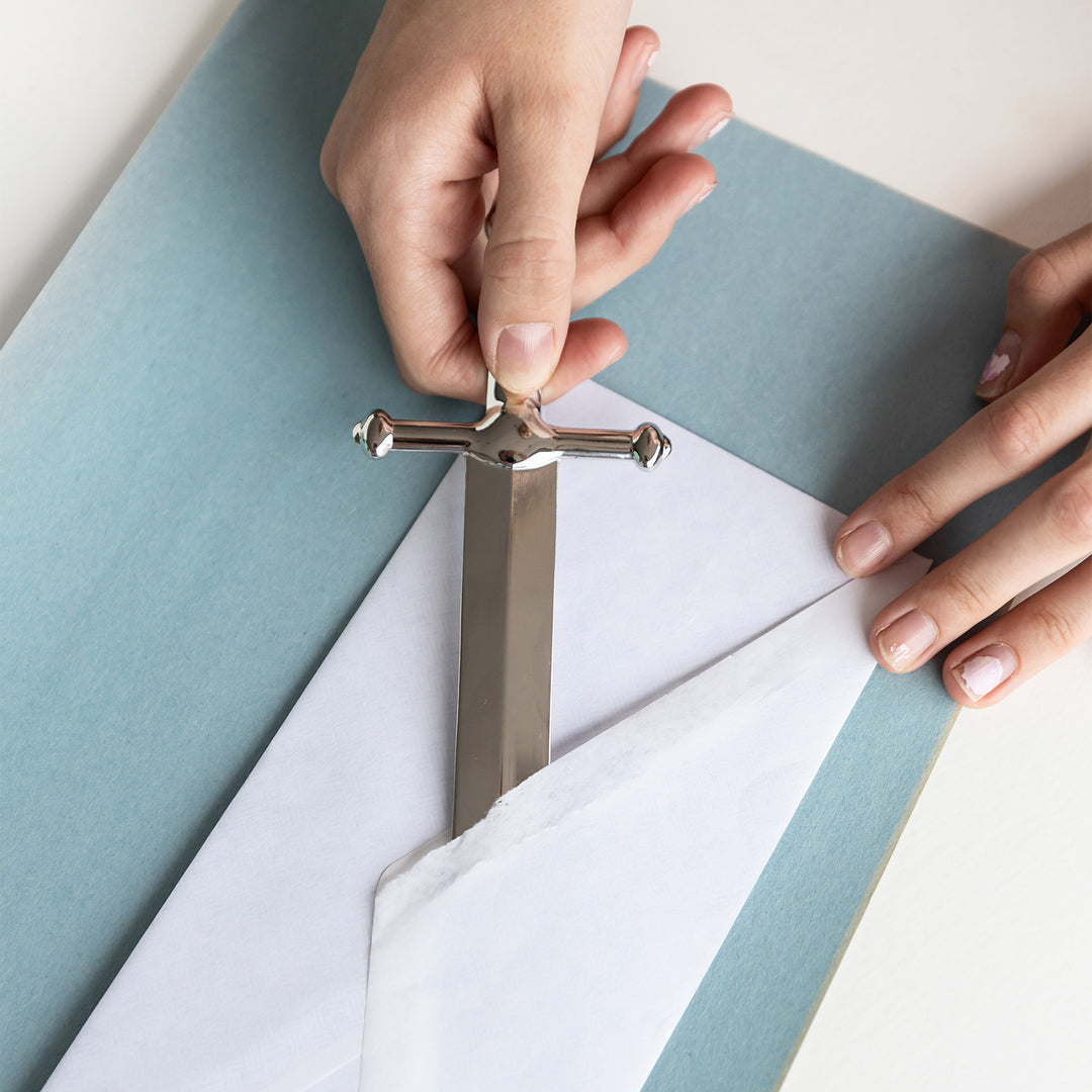 A hand holds a dagger-shaped letter opener, using it to open a white envelope.