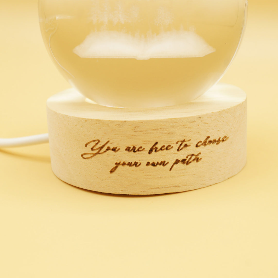 a glass globe light featuring an open book with trees engraved inside the orb. There is a wooden base burned with the quote 'You are free to choose your own path'