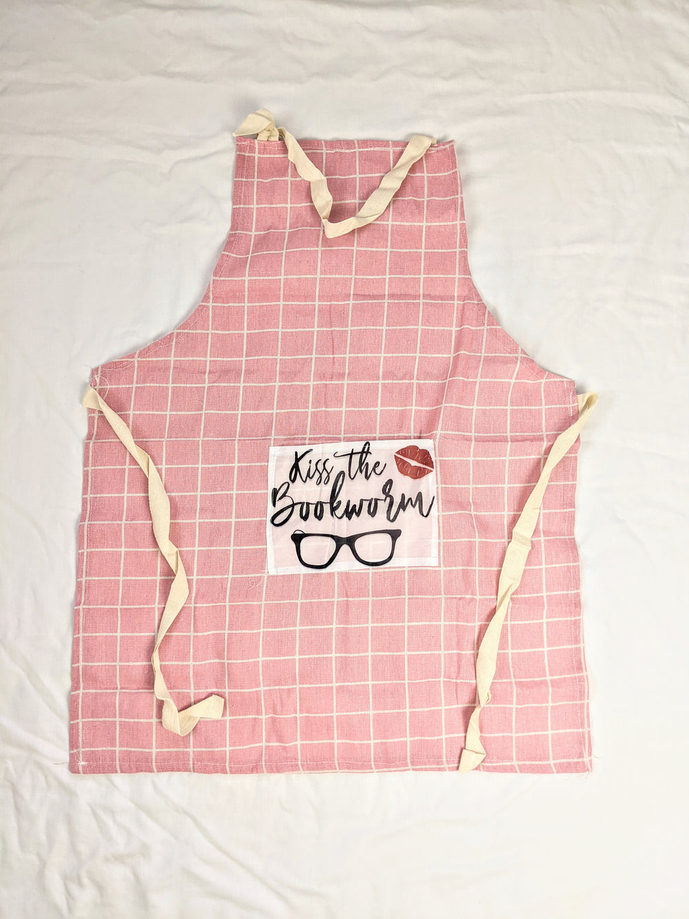 the pink gingham apron featuring a pocket with the words "Kiss the Bookworm" and a pair of glasses and lipstick kiss mark. On a white background.