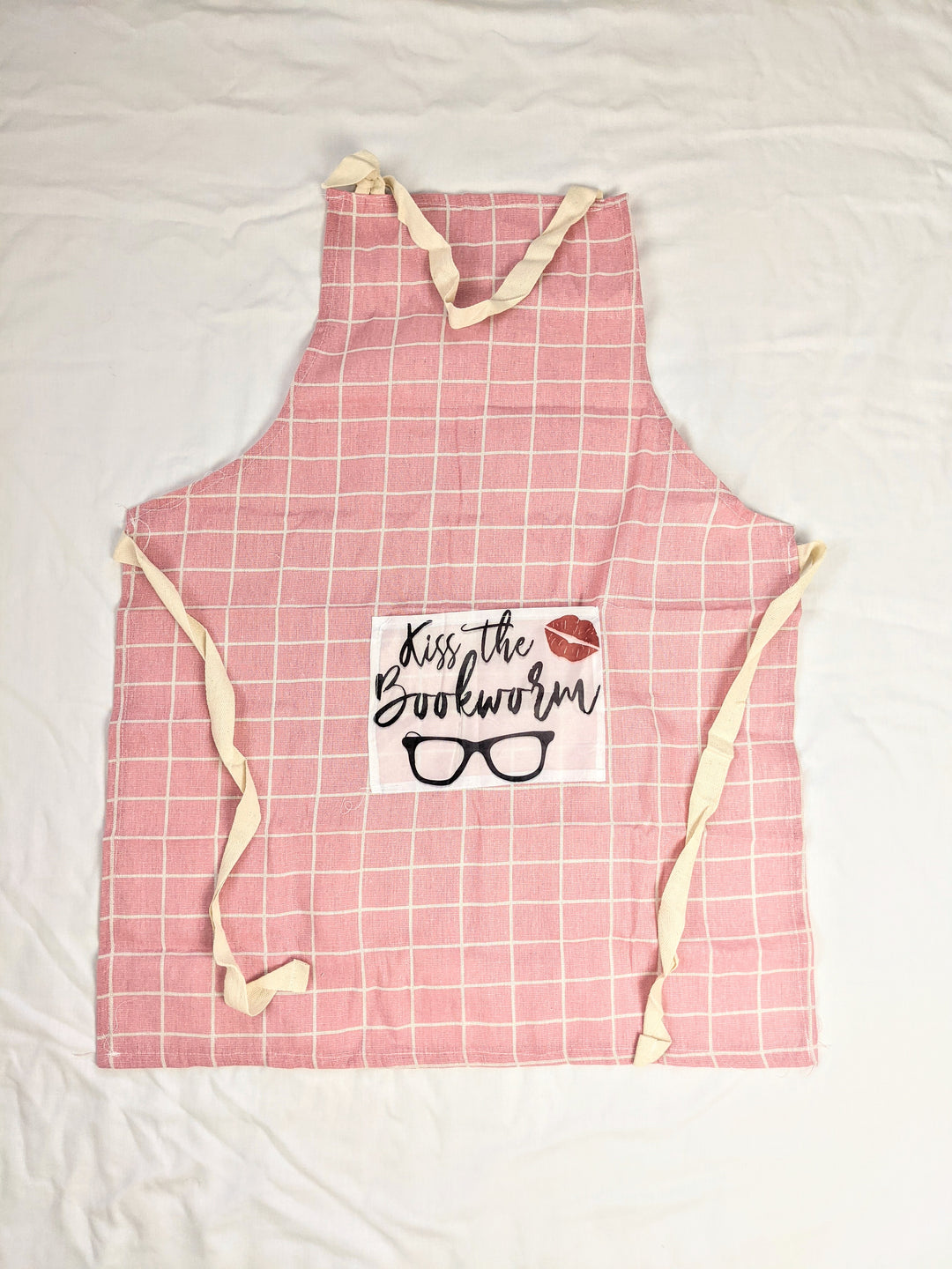 the pink gingham apron featuring a pocket with the words "Kiss the Bookworm" and a pair of glasses and lipstick kiss mark. On a white background.