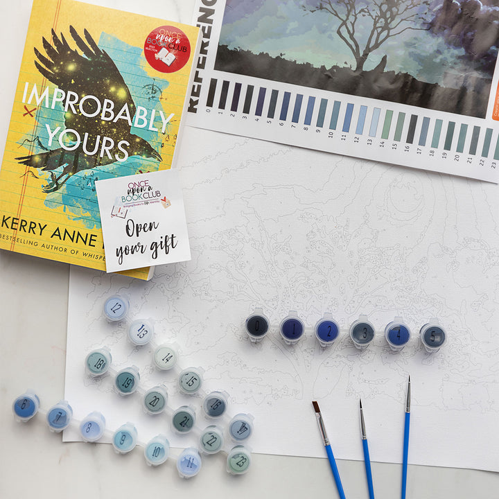 A paint by number kit with a large white paper, multiple sets of paints, reference image of a moonlit tree, 3 paintbrushes, and a paperback edition of Improbably Yours with an open your gift sticker on the front