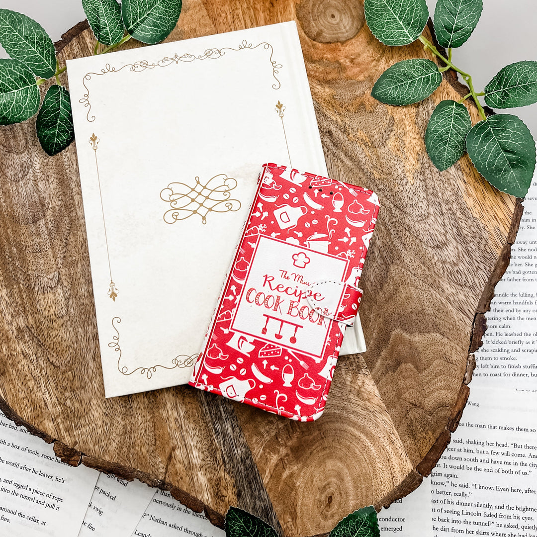 The front of a book-shaped phone case inspired by The Mini Recipe Cook Book. The phone case sits on a white book cover on a wood slice surrounded by greenery.