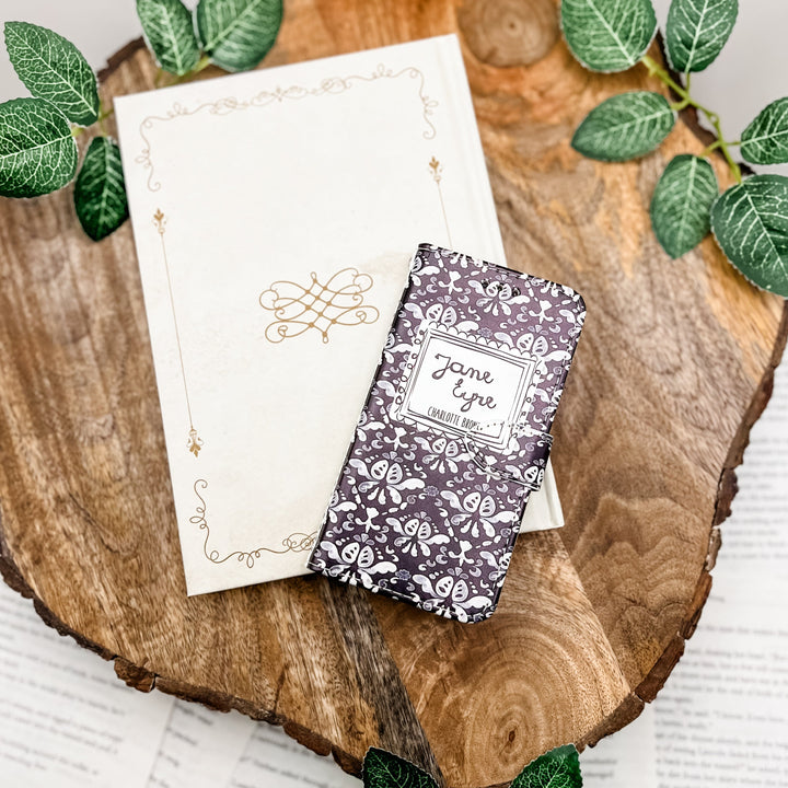 The front cover of a phone case inspired by Jane Eyre by Charlotte Bronte. The phone case sits on a white book on a wood slice surrounded by greenery.