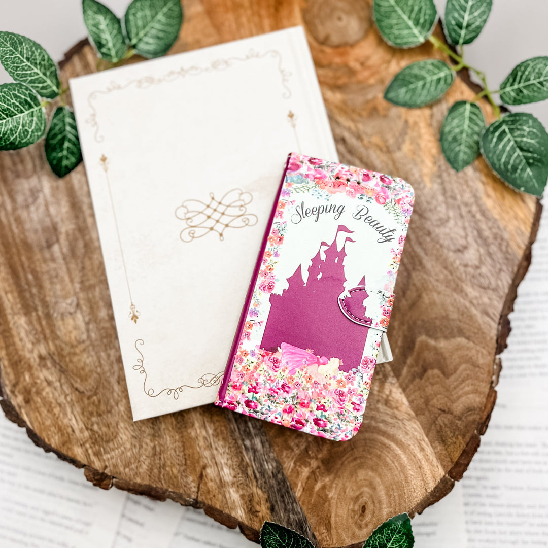 The front cover of a book-shaped phone case inspired by Sleeping Beauty. The phone case sits on a white book cover on top of a wood slice surrounded by greenery.