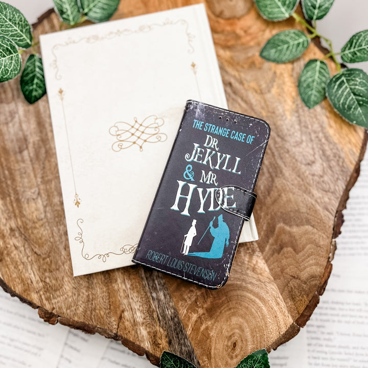 The front cover of a book-shaped phone case inspired by The Strange Case of Dr. Jekyll and Mr. Hyde by Robert Louis Stevenson. The phone case sits on a white book cover on a wood slice surrounded by greenery.