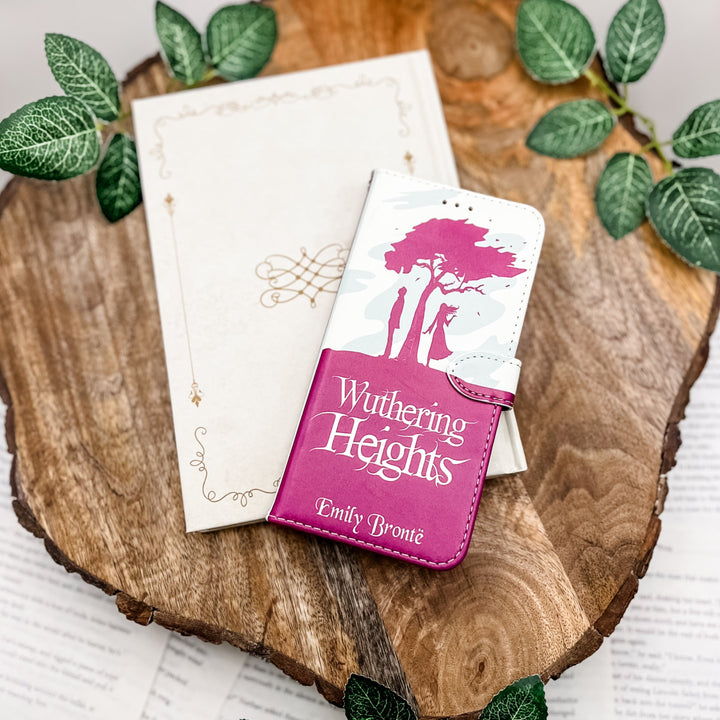 The front of a book-shaped phone case inspired by Wuthering Heights by Emily Bronte. The phone case sits on a white book cover on a wood slice surrounded by greenery.