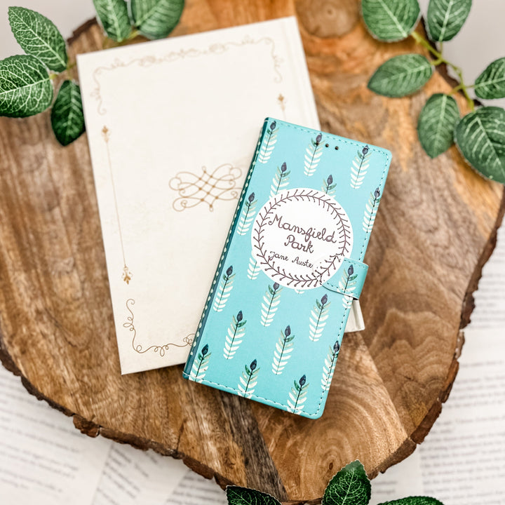 The front of a book-shaped phone case inspired by Mansfield Park by Jane Austen. The phone case sits on a white book cover on a wood slice surrounded by greenery.