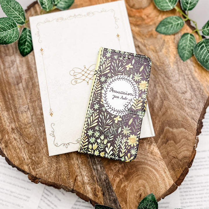 The front cover of a book-shaped phone case inspired by Persuasion by Jane Austen. The phone case sits on a white book cover on a slice of wood surrounded by greenery.