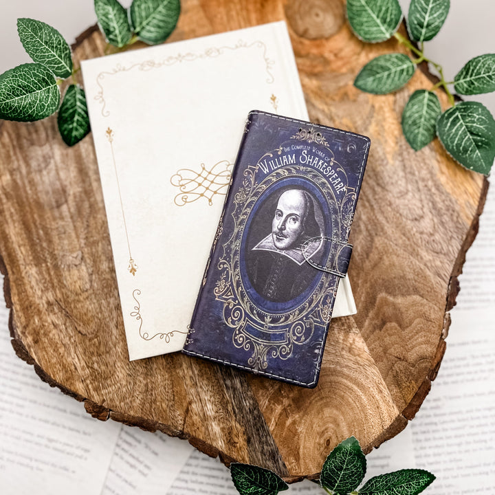 The front cover of a book-shaped phone case inspired by The Complete Works of William Shakespeare. The phone case sits on a white book cover on a slice of wood surrounded by greenery.