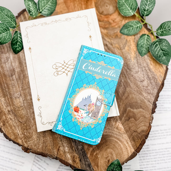 The front cover of a phone case inspired by Cinderella. The phone case sits on a closed white book on a wood slice surrounded by greenery.