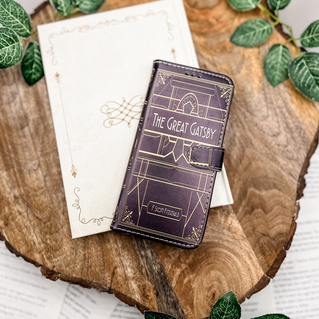 The front of a book-shaped phone case inspired by The Great Gatsby by F. Scott Fitzgerald. The phone case sits on a white book cover on a wood slice surrounded by greenery.