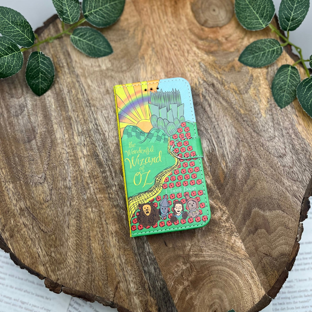 The front cover of a book-shaped phone case inspired by The Wonderful Wizard of Oz by L. Frank Baum. The phone case sits on a wood slice surrounded by greenery.