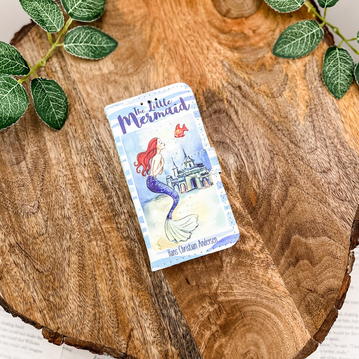 The front of a book-shaped phone case inspired by The Little Mermaid by Hans Christian Andersen. The phone case sits on a wood slice surrounded by greenery.