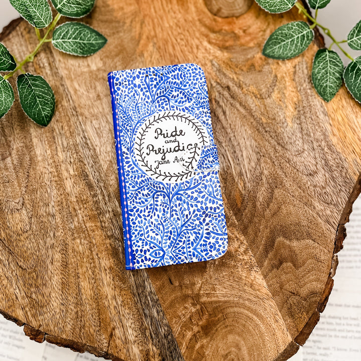 The front cover of a book-shaped phone case inspired by Pride and Prejudice by Jane Austen. The case sits on a wood slice surrounded by greenery.