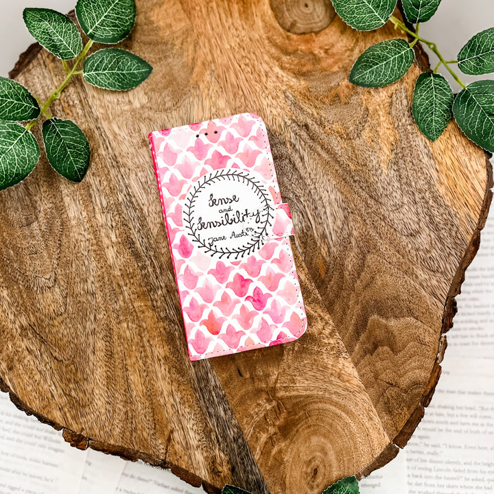 The front cover of a book-shaped phone case inspired by Sense and Sensibility by Jane Austen. The phone case sits on a wood slice surrounded by greenery.