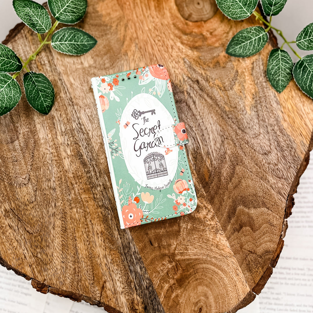 The front cover of a book-shaped phone case inspired by The Secret Garden by Frances Hodgson Burnett. The phone case sits on a wood slice surrounded by greenery.