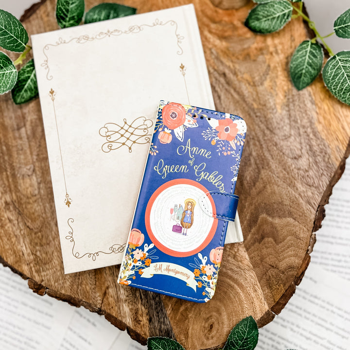 The front cover of a phone case inspired by Anne of Green Gables by L.M. Montgomery. The phone case sits on a white book on a wood slice surrounded by greenery.