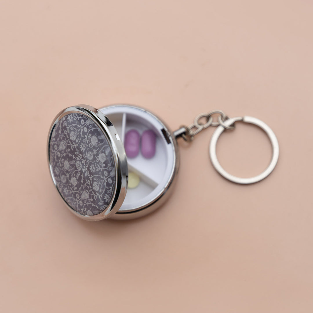 An open pill case on a silver keychain with a purple floral pattern on the front and three separate sections inside.