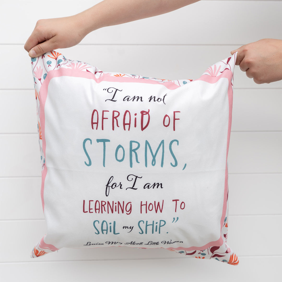 Two hands hold up a square pillow. The pillow is white with a light pink border with floral pattern on the edges. The quote in the middle says I am not afraid of storms, for I am learning how to sail my ship. Louisa May Alcott,  Little Women
