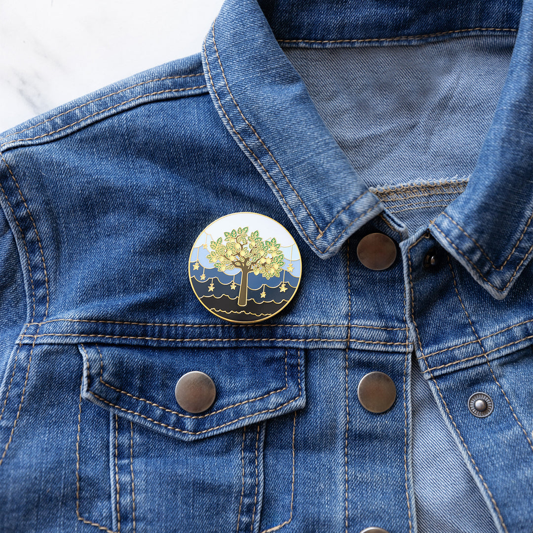 an enamel pin featuring a lemon tree with a sea of stars and clouds behind the tree. pinned to a denim jacket.