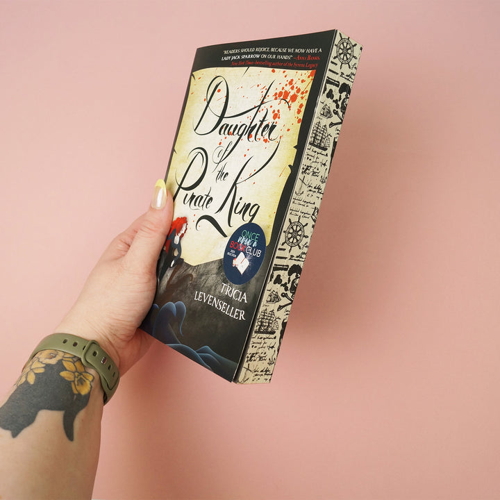 Book titled 'Daughter of the Pirate King' by Tricia Levenseller showcasing patterned page edges is held by a hand on a pink background