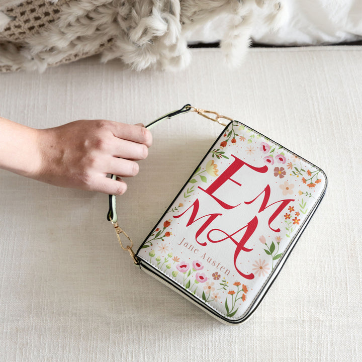 A book-shaped purse inspired by Emma by Jane Austen. A white hand grips the short handle to show its length. The front cover of the purse is visible on a cream-toned, fabric background.