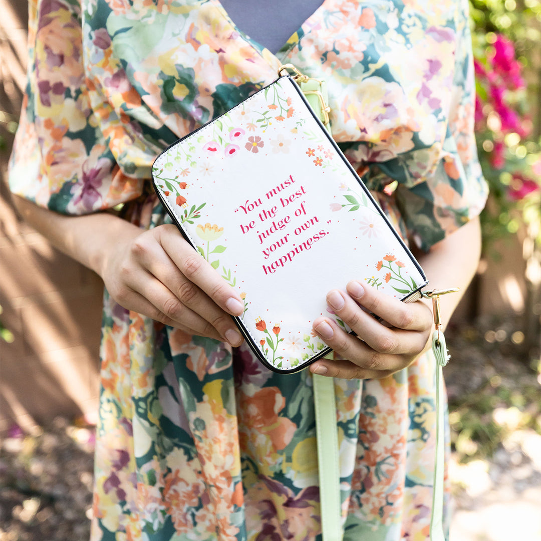 A book-shaped purse inspired by Emma by Jane Austen. A pair of white hands holds the purse to show the back cover with the quote "You must be the best judge of your own happiness" and floral detailing.