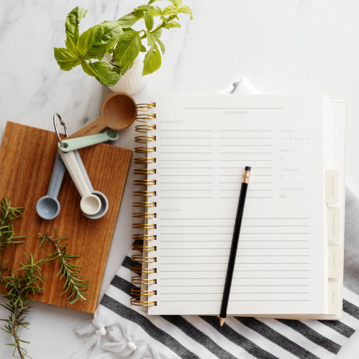 A recipe book lays on a kitchen towel and is open with lined paper and a pencil on top of it. Next to the recipe book are a plant and set of measuring spoons