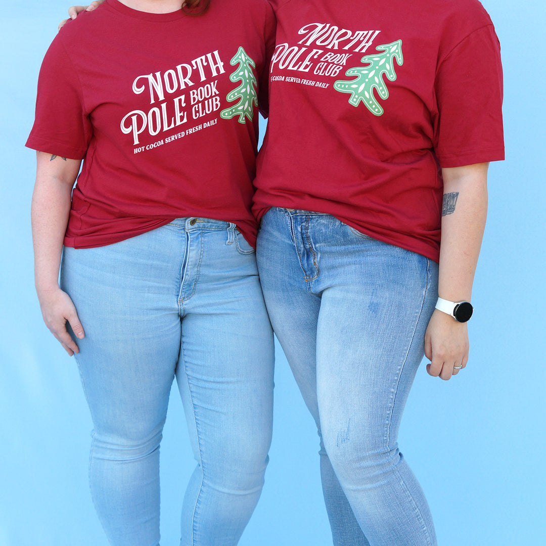Two people wearing red 'North Pole Book Club' t-shirts against a blue background