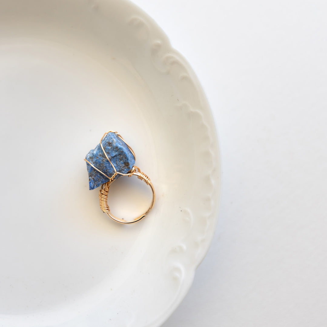 A gold ring with a blue and gray stone laying on a white plate.