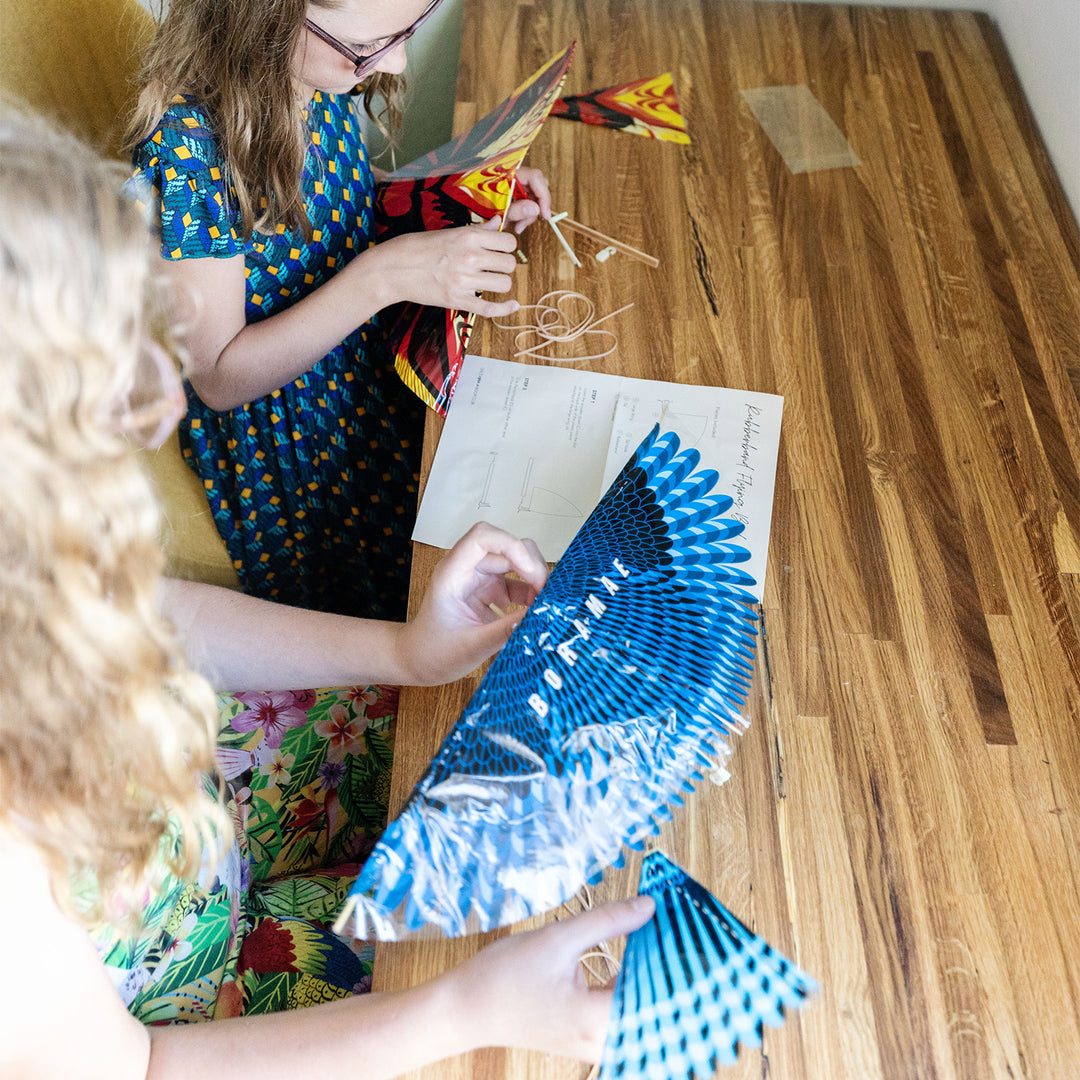 two white children hold rubber-band flying birds, one blue and white and one red and yellow, on a long wooden table