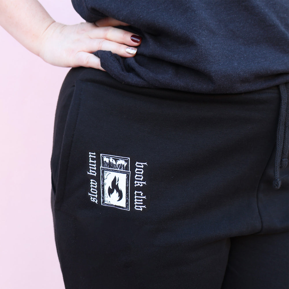 Black sweatpants with embroidered 'Slow Burn Book Club' and matchbook design on a pink background