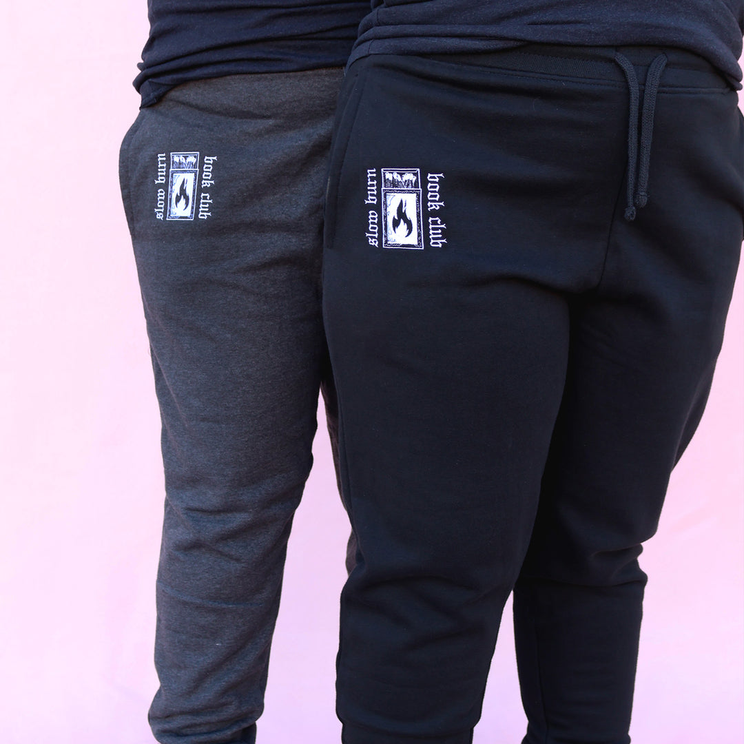 Gray and black sweatpants with embroidered 'Slow Burn Book Club' and matchbook design on a pink background