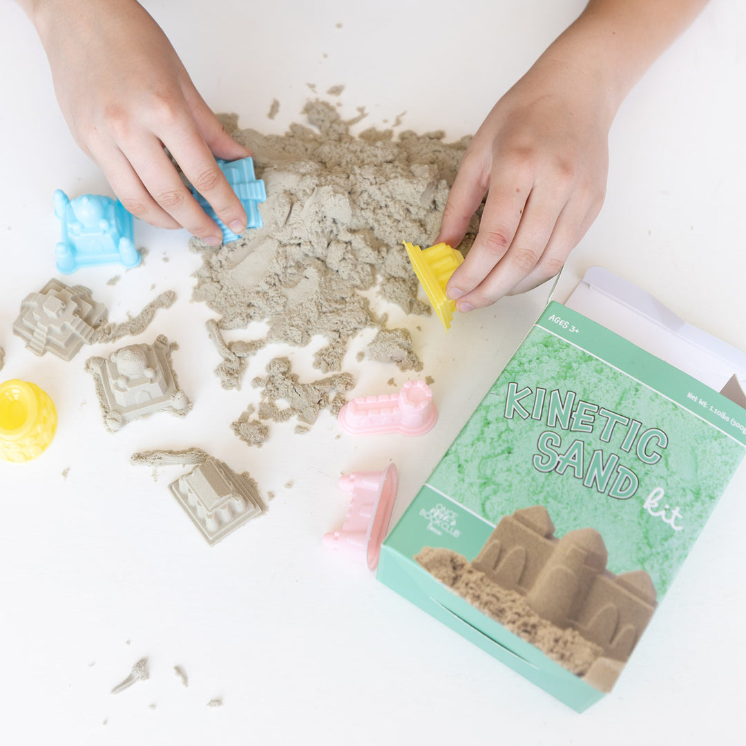 Two hands use multi-colored toys to mold Kinetic sand into castle shapes next to a box labeled Kinetic Sand kit.