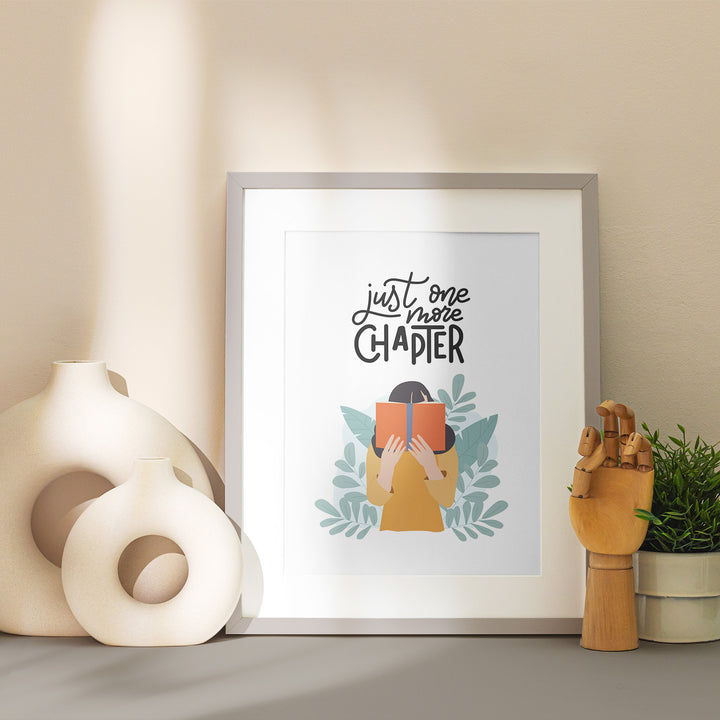 A grey matted frame sits on a table showcasing the "One More Chapter" art print. Around the frame is stylized with vases and home decor in neutral tones. 