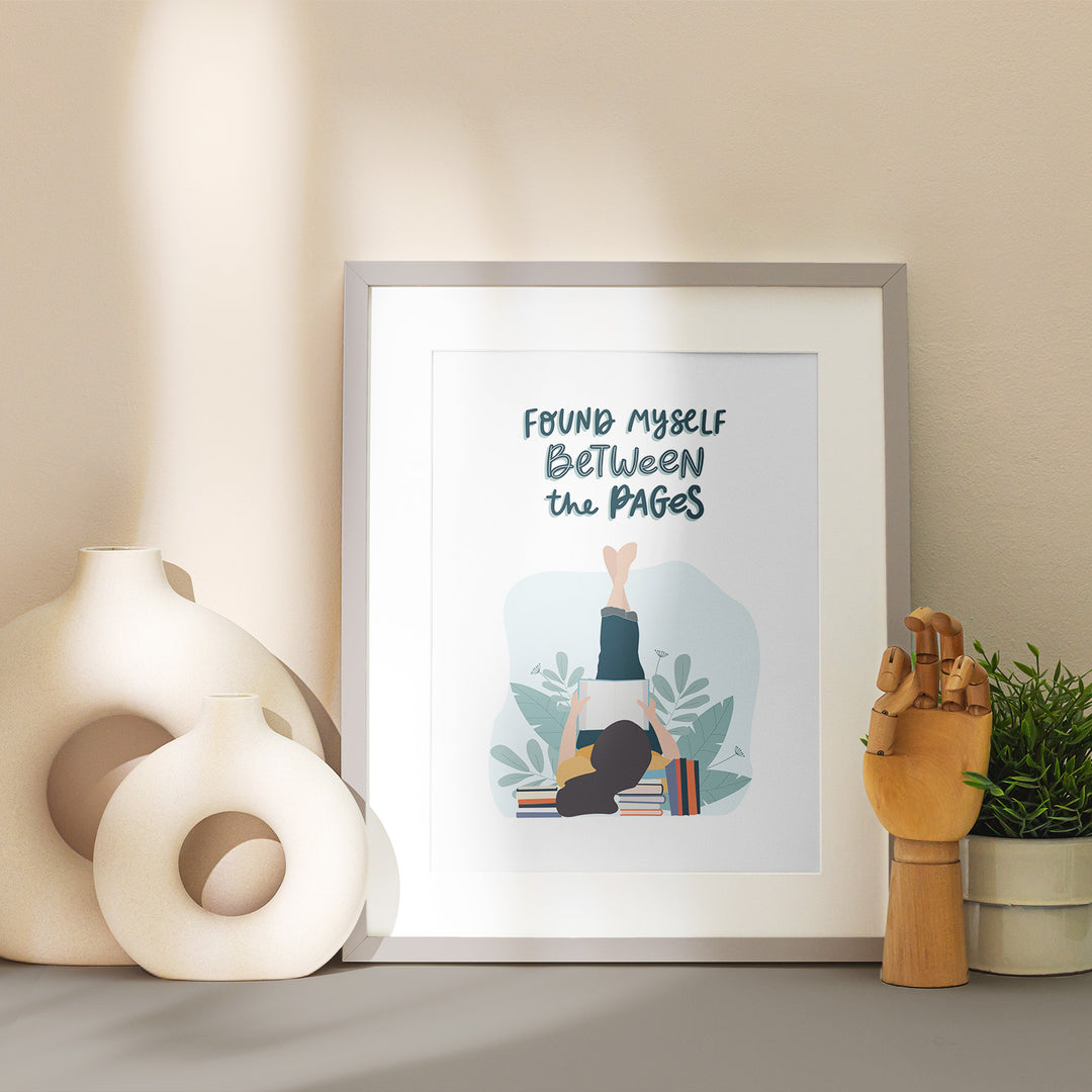 A grey matted frame sits on a table showcasing the "Between the Pages" art print. Around the frame is stylized with vases and home decor in neutral tones.