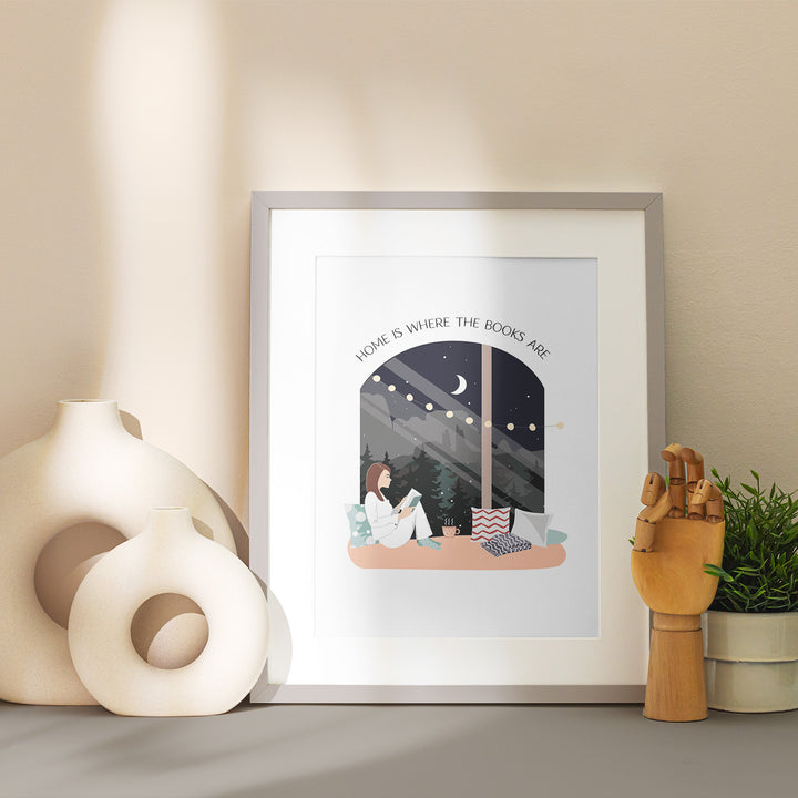 A grey matted frame sits on a table showcasing the "Books Are Home" art print. Around the frame is stylized with vases and home decor in neutral tones.