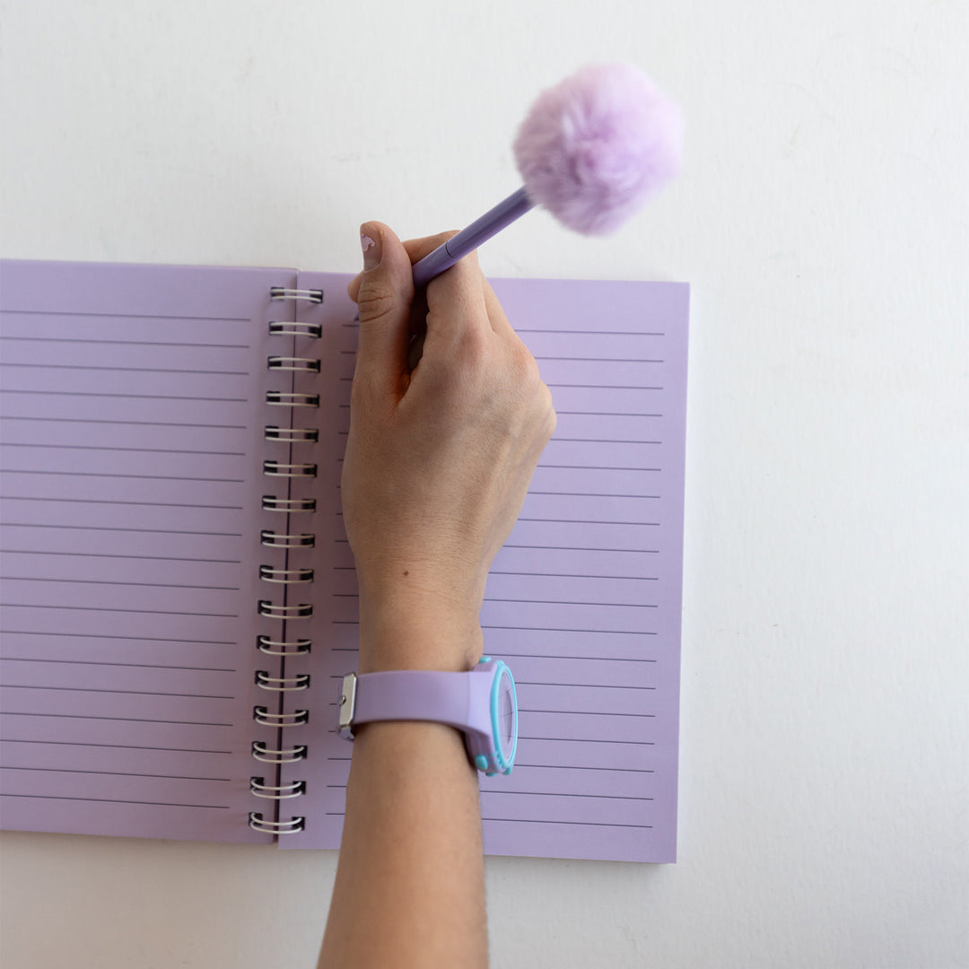 An open light purple notebook with purple pages. A hand lays across the page, holding a purple pen with a matching pom-pom on top.